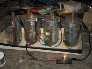 A row of homemade batteries: jam jars and copper pipe and zinc, wired in series on a workbench, one with a little bulb glowing dimly beside it.