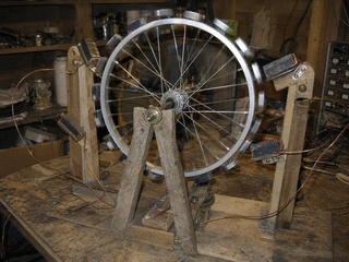 A homemade perpetual-motion wheel on a workbench: a bicycle wheel rim studded with magnets, on a stand, with fixed magnets around it, slightly blurred as if turning.
