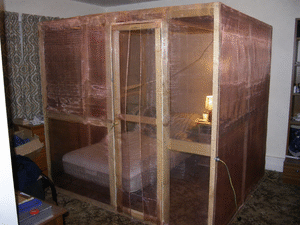 A homemade walk-in Faraday cage in a small spare room: fine copper mesh over a timber frame, a bonded mesh door, an earth wire to a stake, a mattress and a lamp inside.