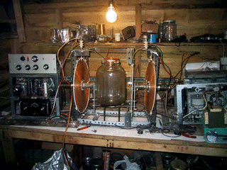 A homemade cyclotron rig on a cluttered shed workbench: a flat copper coil ring, a glass vacuum jar, a rack of dials and oscillators, wires everywhere, lit by a bare bulb.